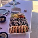 A rectangular plate with oysters, prawns, lemon wedges, and salmon sashimi sits on a white tablecloth with a place setting and a small vase of flowers.