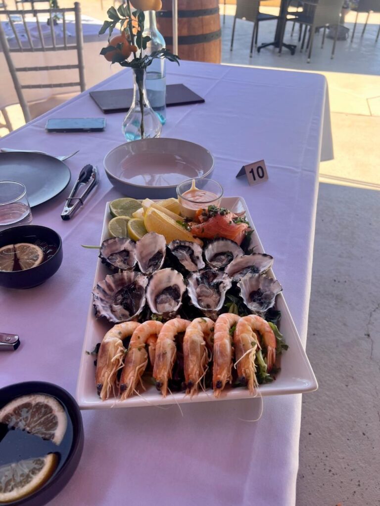 A rectangular plate with oysters, prawns, lemon wedges, and salmon sashimi sits on a white tablecloth with a place setting and a small vase of flowers.