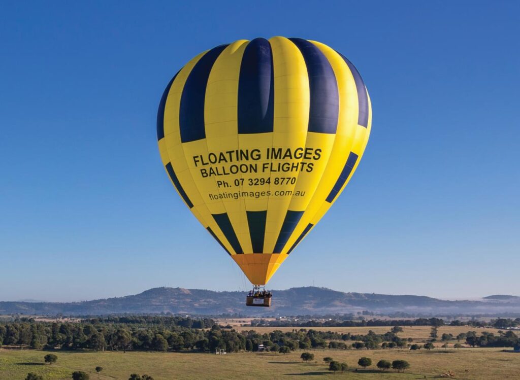 A yellow and blue hot air balloon labeled "FLOATING IMAGES BALLOON FLIGHTS" floats above a rural landscape with hills and trees in the background.