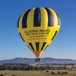 A yellow and blue hot air balloon labeled "FLOATING IMAGES BALLOON FLIGHTS" floats above a rural landscape with hills and trees in the background.
