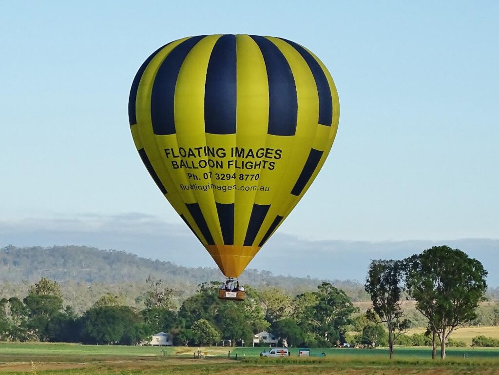 A yellow and black hot air balloon with "Floating Images Balloon Flights" floats above a grassy field with trees and hills in the background.