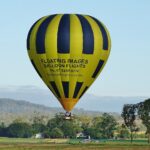 A yellow and black hot air balloon with "Floating Images Balloon Flights" floats above a grassy field with trees and hills in the background.