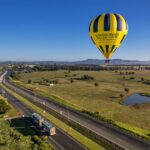 A yellow and black hot air balloon floats above a rural landscape with fields, a pond, and a highway under a clear blue sky.