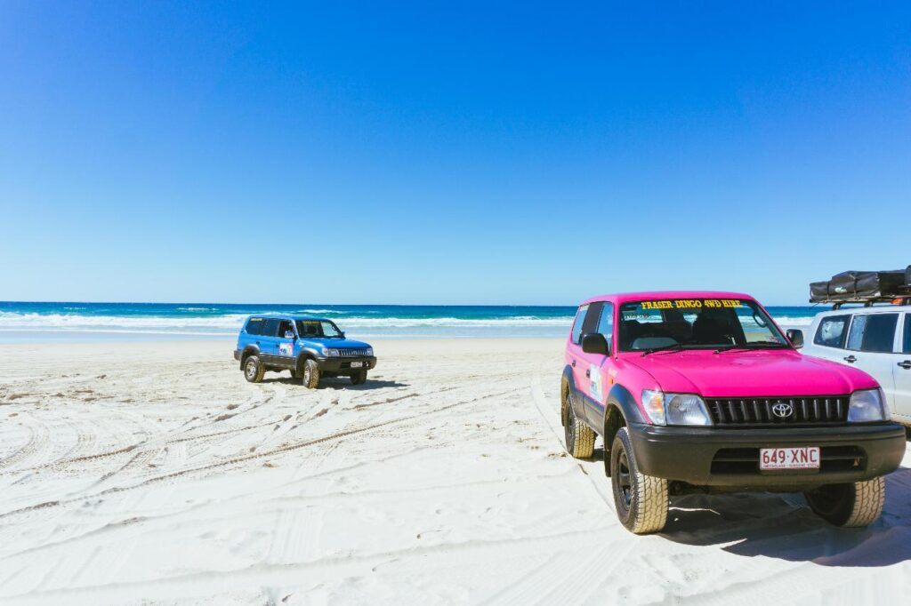 Two four-wheel drive vehicles, one pink and one blue, parked on a sandy beach with the ocean and clear blue sky in the background.