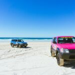 Two four-wheel drive vehicles, one pink and one blue, parked on a sandy beach with the ocean and clear blue sky in the background.