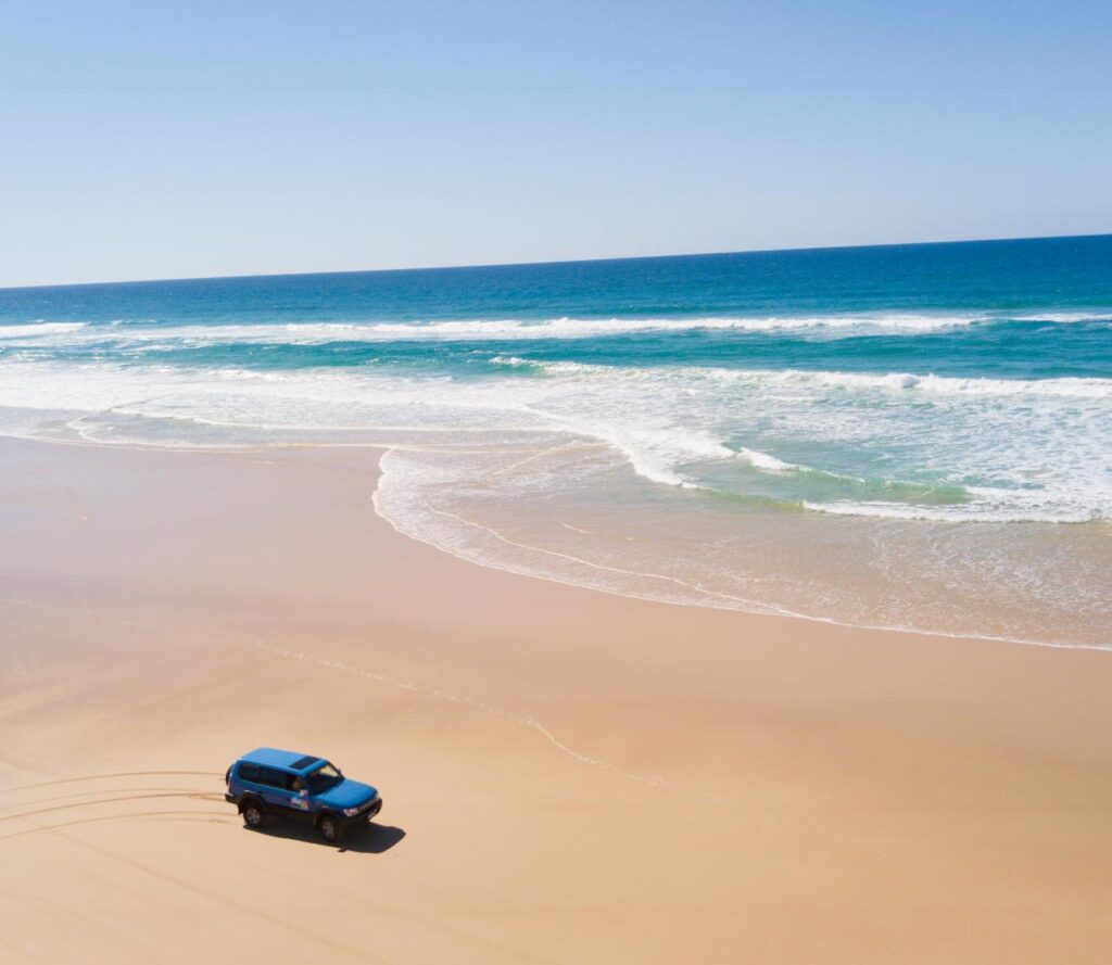 A blue SUV drives along a wide, sandy beach near the shoreline with waves breaking under a clear blue sky.