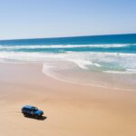 A blue SUV drives along a wide, sandy beach near the shoreline with waves breaking under a clear blue sky.