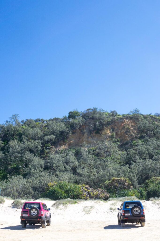 Two SUVs are parked on sandy ground in front of a green, shrub-covered hillside under a clear blue sky.