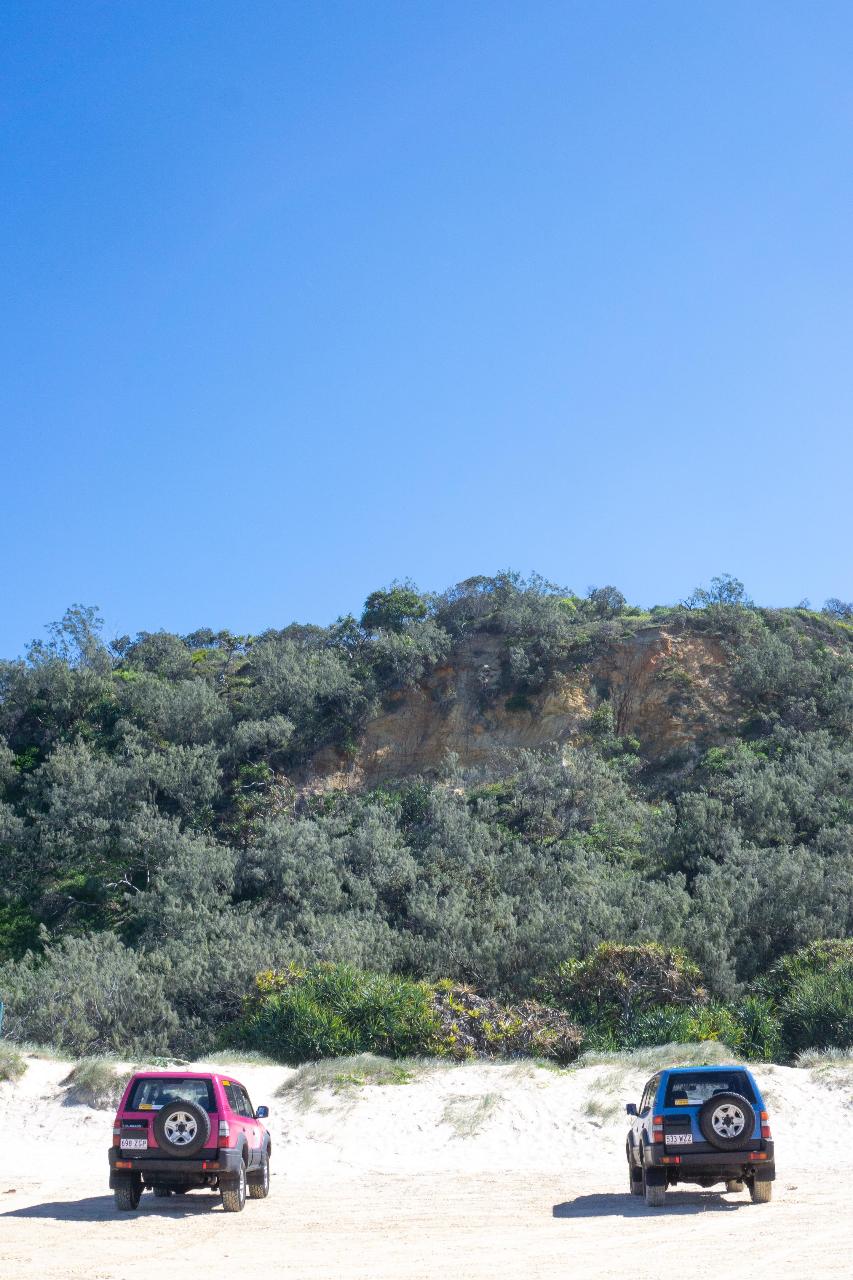 Two SUVs are parked on sandy ground in front of a green, shrub-covered hillside under a clear blue sky.