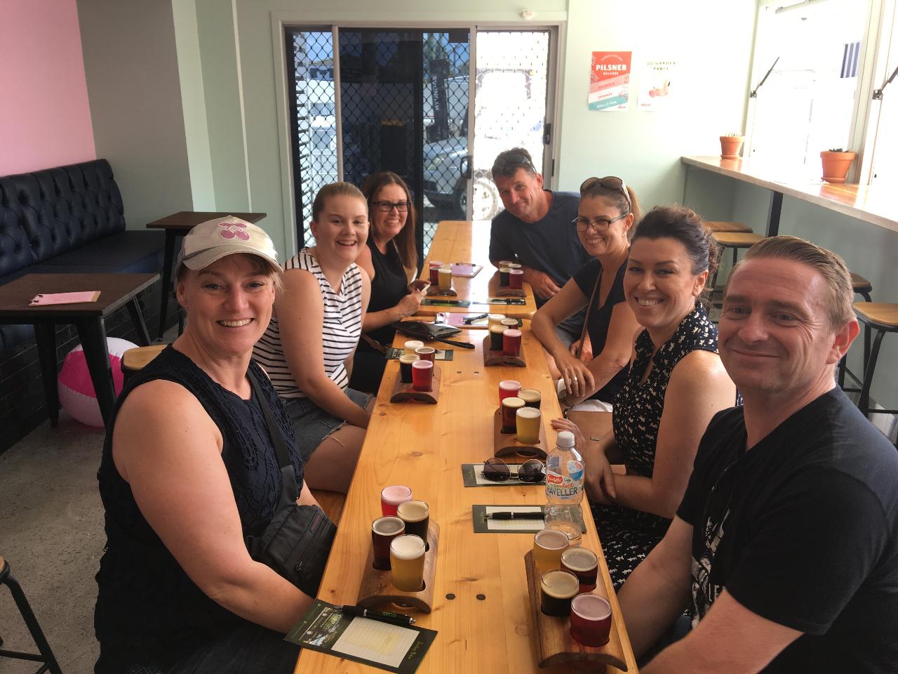 Seven people sit around a wooden table with tasting flights of beer in a casual indoor setting, smiling at the camera.