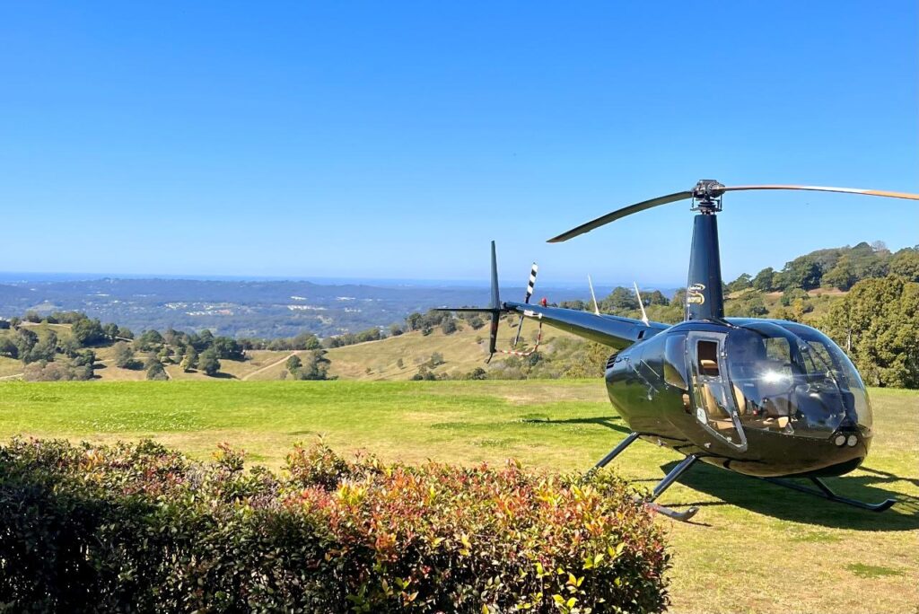 A black helicopter is parked on a grassy field with hills, trees, and a distant landscape under a clear blue sky in the background.
