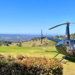 A black helicopter is parked on a grassy field with hills, trees, and a distant landscape under a clear blue sky in the background.