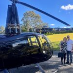 A man and a woman stand next to a black helicopter on a sunny day, with a green lawn, trees, and a building in the background.