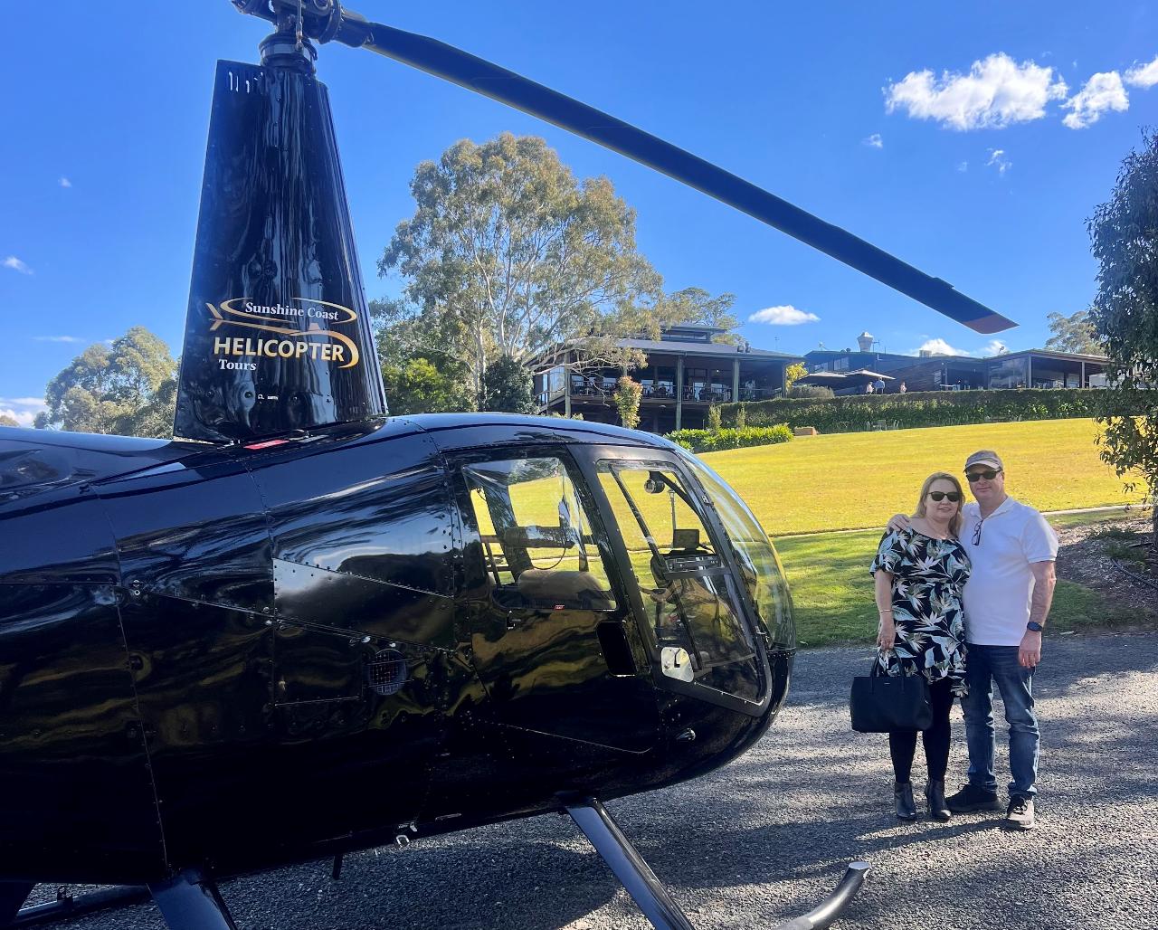 A man and a woman stand next to a black helicopter on a sunny day, with a green lawn, trees, and a building in the background.