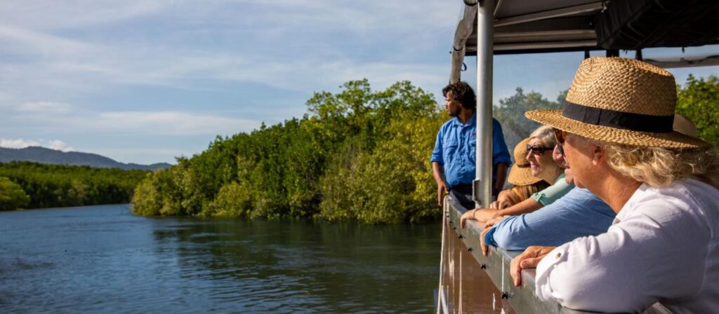Four people on a boat wearing hats look out over a river surrounded by green trees and foliage under a clear sky.