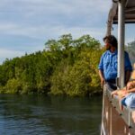 Four people on a boat wearing hats look out over a river surrounded by green trees and foliage under a clear sky.
