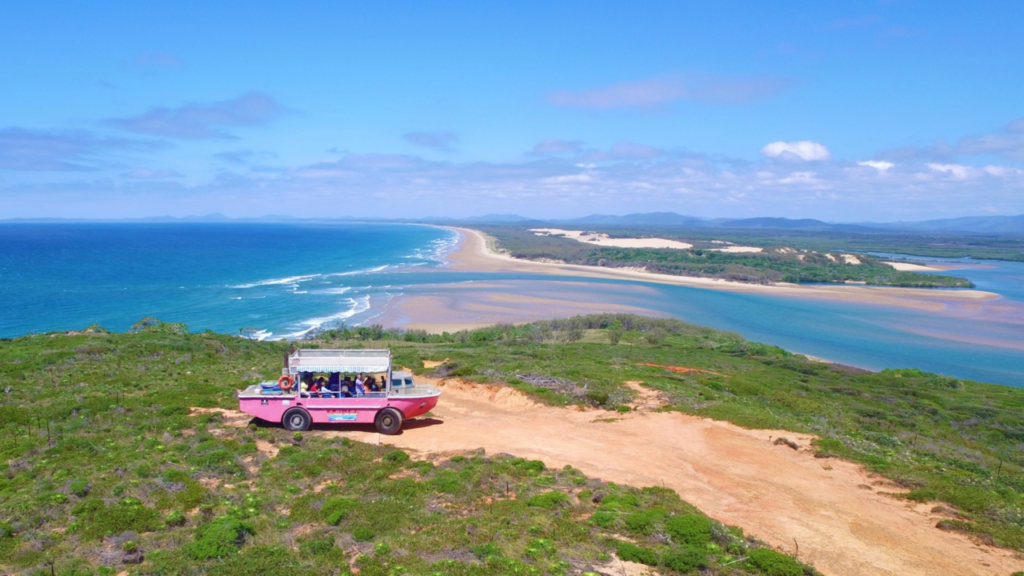 A pink off-road tour vehicle with passengers drives along a dirt path overlooking a scenic coastal landscape with blue ocean, sandy beaches, and green vegetation under a clear sky.