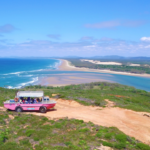 A pink off-road tour vehicle with passengers drives along a dirt path overlooking a scenic coastal landscape with blue ocean, sandy beaches, and green vegetation under a clear sky.
