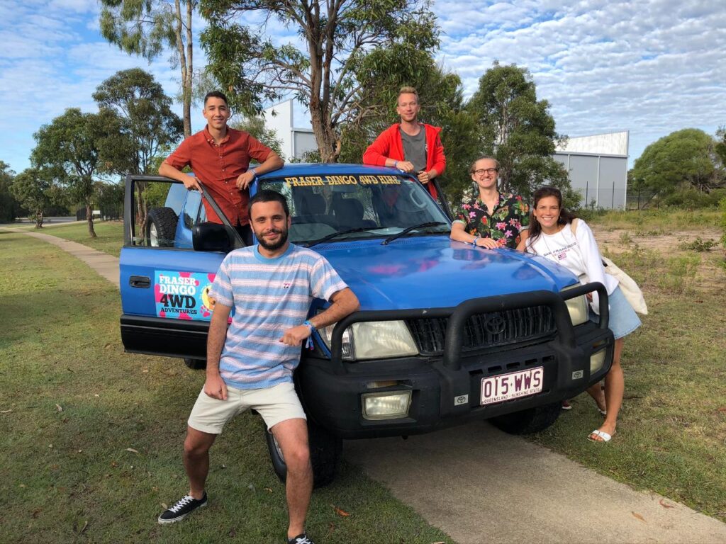 Five people pose in front of a blue 4WD vehicle labeled "Fraser Dingo 4WD Hire" on a paved path with trees and grass in the background.