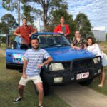 Five people pose in front of a blue 4WD vehicle labeled "Fraser Dingo 4WD Hire" on a paved path with trees and grass in the background.
