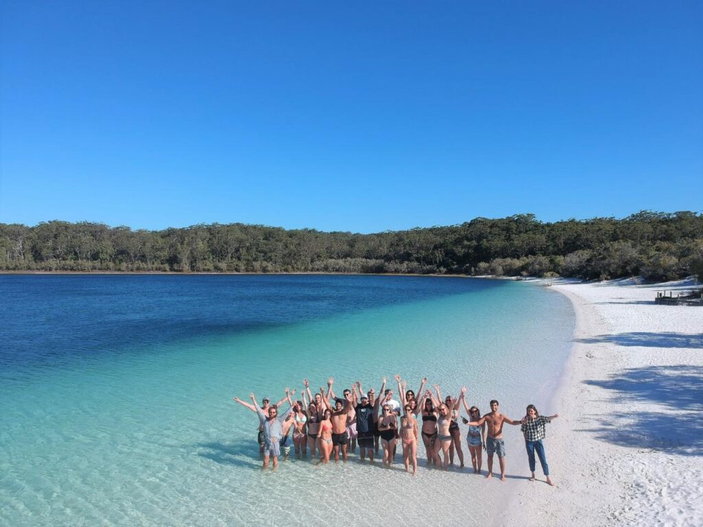 A group of people stand in shallow water and on white sand at the edge of a blue lake with forest in the background under a clear sky.