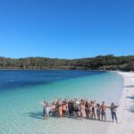 A group of people stand in shallow water and on white sand at the edge of a blue lake with forest in the background under a clear sky.