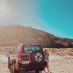 A woman stands next to a red SUV parked on sandy terrain, with grassy hills and a clear blue sky in the background.