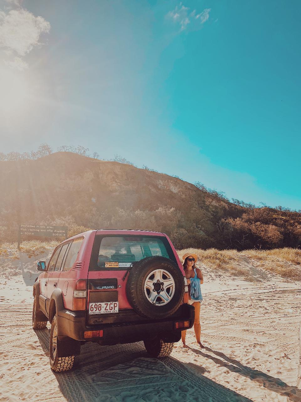 A woman stands next to a red SUV parked on sandy terrain, with grassy hills and a clear blue sky in the background.