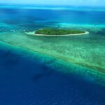 A small, green, tree-covered island surrounded by turquoise and deep blue ocean waters with visible coral reefs.