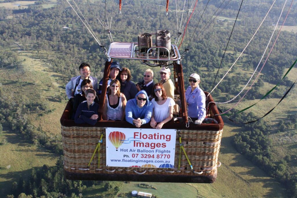 A group of people rides in a hot air balloon basket above a green landscape, with a sign for “Floating Images Hot Air Balloon Flights” attached to the basket.