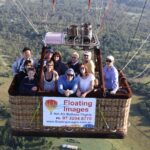 A group of people rides in a hot air balloon basket above a green landscape, with a sign for “Floating Images Hot Air Balloon Flights” attached to the basket.