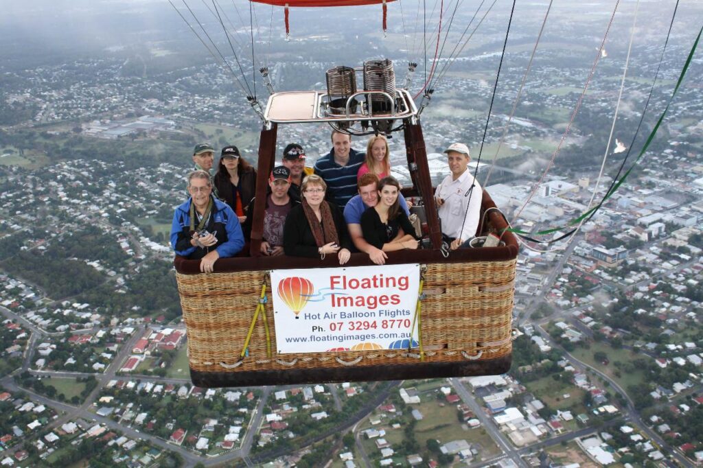 A group of people stand inside a hot air balloon basket in flight above a town, with a banner reading "Floating Images Hot Air Balloon Flights.