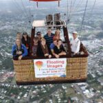 A group of people stand inside a hot air balloon basket in flight above a town, with a banner reading "Floating Images Hot Air Balloon Flights.