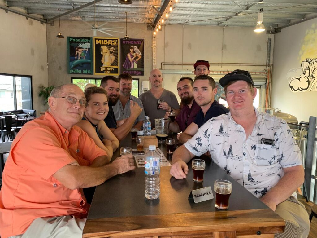 A group of ten people are seated around a table in a bar or brewery, each with drinks in front of them. Posters for different beers hang on the wall in the background.