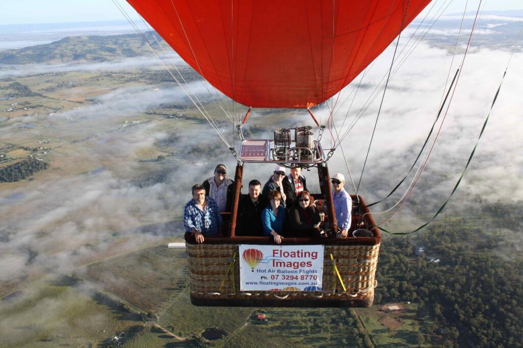 A group of people ride in a hot air balloon basket above a rural landscape, with a banner reading "Floating Images Hot Air Balloon Flights" attached to the basket.
