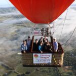 A group of people ride in a hot air balloon basket above a rural landscape, with a banner reading "Floating Images Hot Air Balloon Flights" attached to the basket.