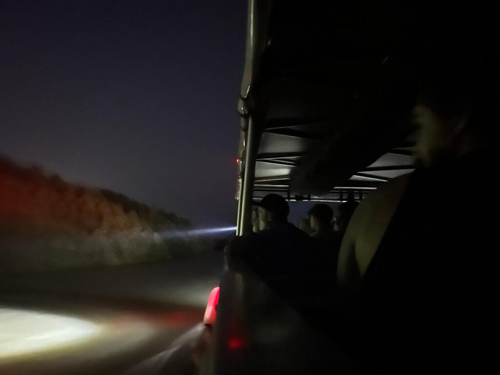 People riding in an open-sided vehicle at night, looking out into the dark with headlights illuminating the road ahead.