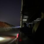 People riding in an open-sided vehicle at night, looking out into the dark with headlights illuminating the road ahead.