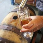 A person fills a glass with brown liquid from a spigot inserted into a wooden barrel, standing on a concrete floor.