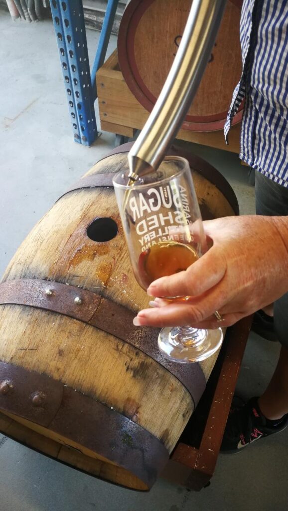 A person fills a glass with brown liquid from a spigot inserted into a wooden barrel, standing on a concrete floor.