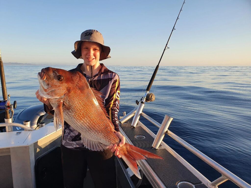Person standing on a boat holding a large fish with one hand, fishing rod nearby, calm sea and clear sky in the background.