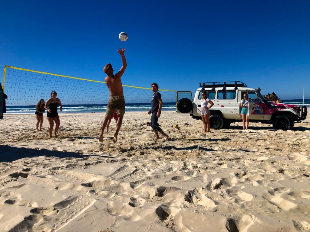 A group of people play volleyball on a sandy beach near the ocean, with an off-road vehicle parked nearby under a clear blue sky.