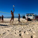 A group of people play volleyball on a sandy beach near the ocean, with an off-road vehicle parked nearby under a clear blue sky.