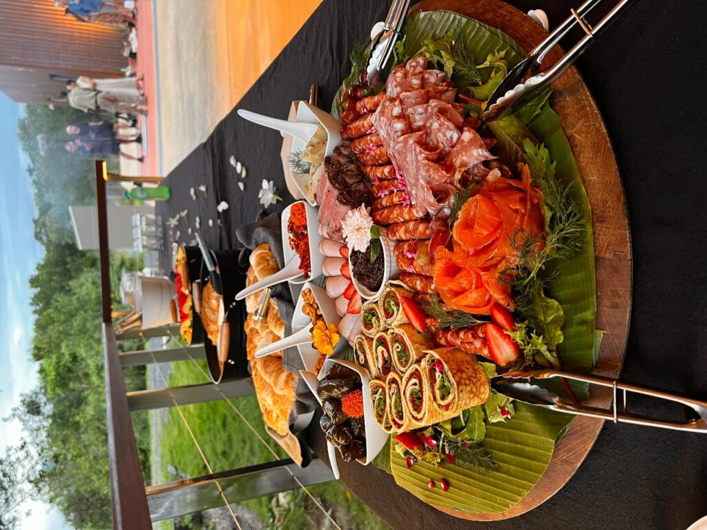 A platter of assorted seafood, meats, and hors d’oeuvres is displayed on a table outdoors, with other dishes arranged behind it.