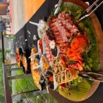 A platter of assorted seafood, meats, and hors d’oeuvres is displayed on a table outdoors, with other dishes arranged behind it.