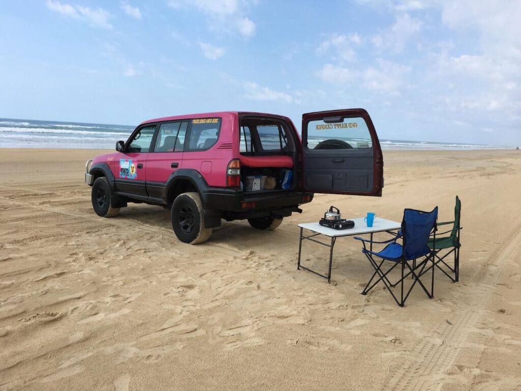 A pink SUV with its rear door open is parked on a sandy beach next to a folding table, camping stove, blue cup, and a blue folding chair. The ocean and sky are in the background.
