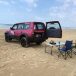 A pink SUV with its rear door open is parked on a sandy beach next to a folding table, camping stove, blue cup, and a blue folding chair. The ocean and sky are in the background.