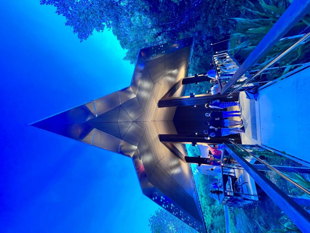 A metallic, arrowhead-shaped structure is illuminated at dusk, with people standing underneath and surrounding trees and a walkway leading up to it.