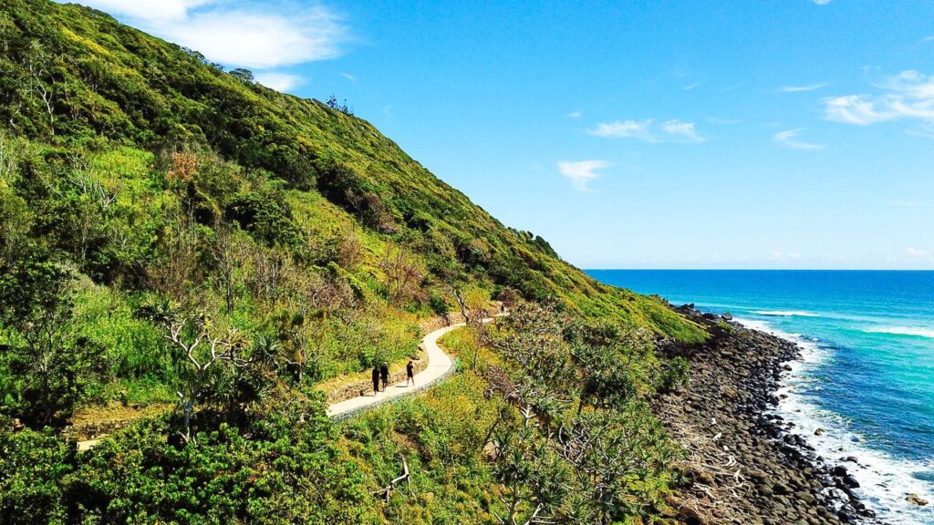 A winding path runs along a green hillside next to rocky shoreline with two people walking under a clear blue sky.
