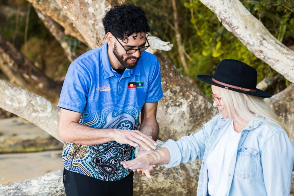 A man in a patterned blue shirt applies ochre paint to a woman’s forearm outdoors, with large tree branches in the background.